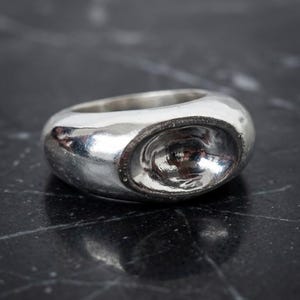 Peut inclure: Une bague épaisse en argent poli avec un anneau lisse et arrondi, présentant une indentation ovale concave marquée sur la face avant. Le métal possède une finition brillante et réfléchissante qui capte la lumière, créant des reflets intenses.