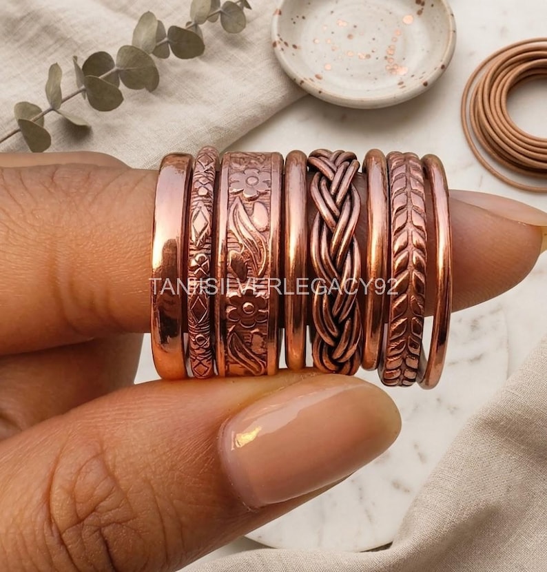 May include: A set of seven copper rings featuring various textures and patterns, including smooth bands, a floral motif, a braided design, and a leaf-like pattern. The text TANISILVERLEGACY92 is visible across the center of the rings.