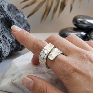 Könnte beinhalten: Ein silberfarbener Ring im offenen Bypass-Design mit einer texturierten, gehämmerten Oberfläche. Das Band besteht aus zwei überlappenden Enden, die sich nicht berühren und eine deutliche Lücke in der Metallstruktur auf dem Finger bilden.