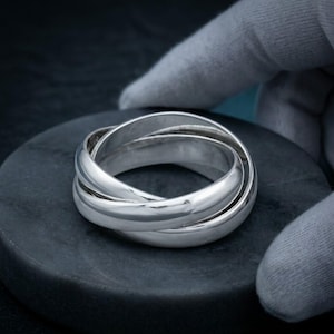 Könnte beinhalten: Ein Ring aus poliertem Silber, der aus drei ineinandergreifenden Bändern besteht, die sich zu einer kreisförmigen Struktur überlappen. Die Metalloberfläche ist glatt und reflektierend und zeigt einen hellen Glanz an den abgerundeten Kanten.