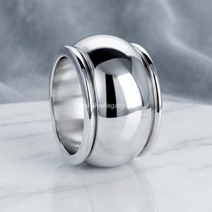 Può includere: Un anello largo in metallo color argento lucido con una superficie convessa liscia e un bordo rialzato prominente su entrambi i lati. Il metallo ha una finitura altamente riflettente, simile a uno specchio, che cattura la luce e crea riflessi.