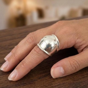 May include: A wide, polished silver ring featuring a prominent, smooth, dome-shaped centrepiece that extends outwards from the band. The metal has a reflective, high-shine finish with a curved, rounded silhouette that sits across the finger.