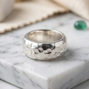 Peut inclure: Une large bague en argent présentant une finition martelée texturée sur toute sa surface extérieure. Le métal a un aspect poli et réfléchissant avec de nombreuses petites indentations arrondies créant un motif distinct et irrégulier.