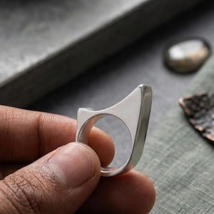 May include: A silver ring with a smooth, polished finish and a distinct, asymmetrical geometric shape. The band features a circular opening for the finger, while the upper portion extends into a pointed, angular silhouette with flat edges.