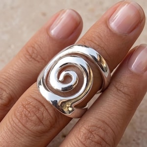 Peut inclure: Une bague en argent poli présentant un motif en spirale ajouré proéminent qui s'enroule autour du doigt. Le métal possède une surface lisse et réfléchissante avec une forme fluide et continue créant un motif circulaire sur l'avant.
