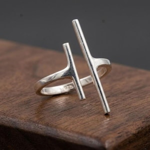 Op de afbeelding: Een open zilveren ring met twee verticale staven van verschillende lengtes die uit de ringband steken. Het metalen oppervlak is gepolijst en reflecterend, met het keurmerk 925 duidelijk gestempeld aan de binnenkant van de ring, wat duidt op sterling zilver.