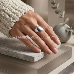 Peut inclure: Une bague en argent poli au design ouvert et asymétrique, dotée d'une surface lisse et incurvée qui s'enroule autour du doigt. La bague présente une finition métallique réfléchissante et un large anneau qui s'affine en pointe. Le texte TANIISILVERLEGACY92 est visible.