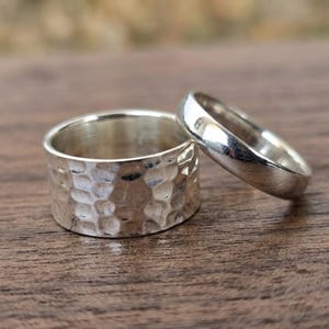 May include: Two silver-colored rings. One ring is wide with a textured, hammered surface finish. The second ring is narrower with a smooth, polished, and reflective surface. Both rings are metallic and have a rounded, circular band shape.