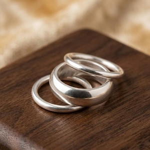 Op de afbeelding: Een set van drie gepolijste zilverkleurige ringen die op elkaar zijn gestapeld. De ringen hebben een glad, afgerond oppervlak met een reflecterende metallic afwerking. De banden variëren enigszins in dikte, wat zorgt voor een gelaagd uiterlijk.