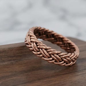 Op de afbeelding: Een koperen ring met een doorlopend gevlochten of geweven metaalpatroon. De ring heeft een warme, metaalachtige roodbruine afwerking met een getextureerd oppervlak dat wordt gevormd door de in elkaar grijpende metalen strengen.