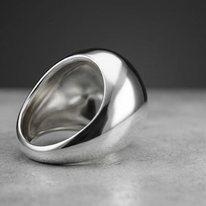 Puede incluir: Un anillo de tono plateado pulido con una banda gruesa, redondeada y lisa. El anillo presenta una superficie superior ancha y abovedada que se estrecha hacia abajo en una forma circular sólida y continua, reflejando la luz en su acabado.