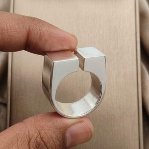 Può includere: Un anello color argento caratterizzato da una fascia larga e liscia con un design aperto sulla parte superiore. La parte superiore dell'anello è composta da due blocchi rettangolari distinti con sommità piatta, separati da un piccolo spazio.