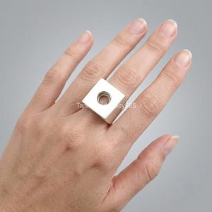 Könnte beinhalten: Ein silberfarbener Ring mit einem markanten, flachen, quadratischen Blockdesign. Die Mitte der quadratischen Oberfläche enthält einen kreisförmigen Ausschnitt oder ein Loch. Der Ring wird an einem Finger getragen und zeigt das metallische Finish und die geometrische Form.