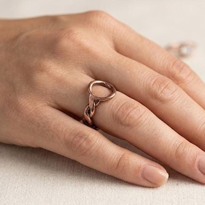 Può includere: Un anello color rame caratterizzato da un elemento circolare superiore collegato a una fascia intrecciata simile a una corda. Il metallo ha una finitura calda con una texture visibile lungo il gambo intrecciato, creando un contrasto tra il cerchio liscio e il design intrecciato.