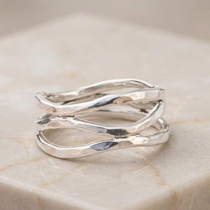 Könnte beinhalten: Ein Silberring mit einem mehrreihigen Design aus drei markanten, wellenförmigen und gehämmerten Metallsträngen, die an den Seiten miteinander verbunden sind. Die Silberoberfläche hat ein glänzendes Finish mit sichtbaren Hammerschlagspuren.