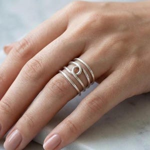 Puede incluir: Un anillo de plata de varias bandas que presenta cuatro bandas finas, redondeadas y distintas que convergen en un punto central. El diseño crea una apariencia apilada con un acabado liso y pulido que envuelve el dedo en un movimiento fluido.
