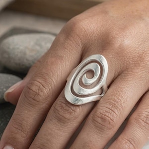 Può includere: Un anello in argento caratterizzato da un grande design a spirale traforato che copre il dito. Il metallo presenta una finitura liscia e lucida con una forma continua e fluida che crea un motivo circolare al centro dell'anello.