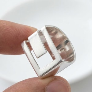 Op de afbeelding: Een zilveren ring met een open, geometrisch vierkant ontwerp en een gladde, gepolijste afwerking. De binnenkant van de ring is duidelijk gestempeld met het getal 925, wat het zilvergehalte van dit sieraad aangeeft.