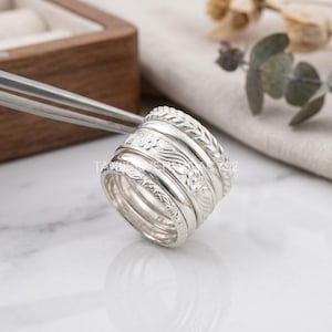 Può includere: Un set di cinque anelli in argento impilati tenuti da una pinzetta metallica. Gli anelli presentano texture varie tra cui un motivo intrecciato, un design floreale con foglie, una fascia liscia lucida e una fine incisione a foglia.