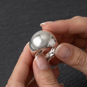 Peut inclure: Une bague argentée polie dotée d'un anneau large, lisse et arrondi en forme de dôme. La surface de la bague est hautement réfléchissante, créant une finition miroir qui capte la lumière sur l'ensemble de son design extérieur bombé.