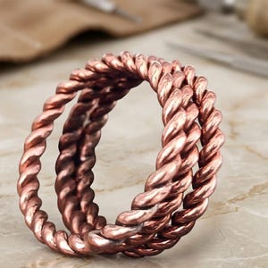 May include: A copper ring featuring a triple-layered design composed of three distinct twisted rope-style bands. The metallic surface exhibits a warm, reddish-brown hue with polished highlights that emphasize the intricate, spiraling texture of the metal.