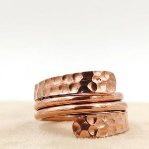 Op de afbeelding: Een koperen ring met een wikkelontwerp bestaande uit drie afzonderlijke banden. De twee buitenste banden hebben een gehamerde, getextureerde afwerking, terwijl de middelste band glad en rond is. Het koper heeft een warme, roodbruine kleur.