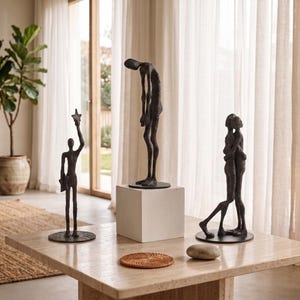 May include: Three dark bronze-toned metal sculptures on a stone table. One figure holds a star, one stands with a slumped posture, and two figures embrace. Each sculpture is mounted on a circular base, with the central figure elevated on a white cube.
