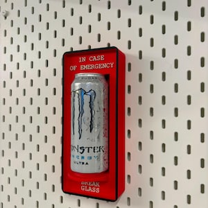 May include: A rectangular red wall-mounted display case containing a silver Monster Energy Ultra can. The case features black trim and white text that reads IN CASE OF EMERGENCY at the top and BREAK GLASS at the bottom, framing the beverage can.
