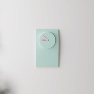 Könnte beinhalten: Eine rechteckige Wanduhr in einem einfarbigen, hellen Mintgrün mit einem kreisförmigen Zifferblattbereich. Die Uhr hat zwei einfache, flache Zeiger in einem zarten Rosaton, die die Zeit anzeigen, ohne dass Zahlen oder Markierungen vorhanden sind.
