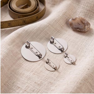 May include: Four circular silver-toned metal brooch blanks with attached pin backs in varying sizes. A yellow measuring tape is visible in the upper left corner, showing markings in inches. The items are arranged on a textured beige fabric surface.