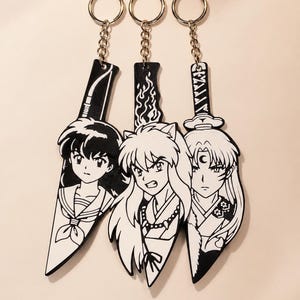 Peut inclure: Trois porte-clés en noir et blanc en forme de lames de couteau, chacun présentant une illustration de personnage d'anime différente. Les motifs incluent une fille aux longs cheveux noirs, un personnage aux oreilles de chien et un croissant de lune.