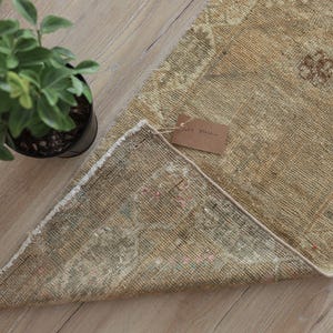 May include: A rectangular woven rug featuring a muted tan and beige color palette with intricate faded floral patterns. The rug is partially folded over to reveal its underside and includes a small brown paper tag attached by a string.
