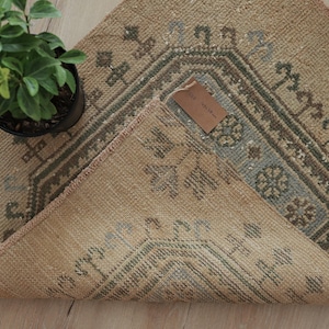 May include: A rectangular rug with a tan base color featuring a geometric pattern in muted blue, green, and brown tones. The rug is folded over at one corner, revealing a brown paper tag with the text 45 x 88 cm printed on its surface.