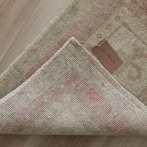 May include: A rectangular rug with a folded corner revealing a woven grid backing and a light beige and faded red patterned front. A small brown rectangular paper tag with illegible handwritten text is attached to the rug with a thin string.