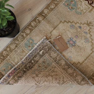 May include: A rectangular rug with a beige base, featuring a repeating pattern of floral motifs in muted blue, pink, and brown tones. The rug is partially folded over to reveal a dark brown underside and has a small brown paper tag attached.