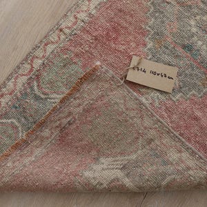 Puede incluir: Una alfombra doblada con un patrón descolorido en tonos rosa viejo, gris apagado y toques de verde azulado. Una pequeña etiqueta de papel beige está adherida con el texto escrito a mano 6714 110x47cm, indicando las dimensiones de la pieza.