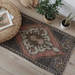 Op de afbeelding: Een rechthoekig vloerkleed met een centraal ruitpatroon in tinten van gedempt roze, antracietgrijs en crème. Het kleed heeft een geometrische rand en een verweerde afwerking, met een herhalend motief over het geweven oppervlak.