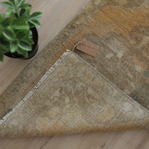 Könnte beinhalten: Ein rechteckiger Teppich mit einem verblassten, abgenutzten Muster in Senfgelb, Olivgrün und Beige. Der Teppich hat an der Kante eine kurze weiße Franse und ein kleines braunes Papieretikett, das mit einer Schnur an der Oberfläche befestigt ist.
