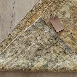 May include: A folded rectangular rug featuring a mustard yellow and tan geometric pattern with a visible woven texture on the underside. A small brown paper tag is attached to the corner of the rug, displaying handwritten text in black ink.