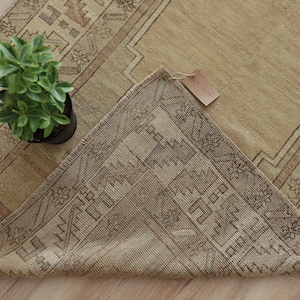 Puede incluir: Una alfombra rectangular con una paleta de colores beige y arena, que presenta patrones geométricos y un diseño de borde. Una esquina de la alfombra está doblada para revelar el reverso, que muestra un patrón similar en tonos marrones apagados.