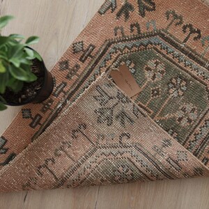 May include: A rectangular woven rug featuring a muted orange background with a central green medallion and dark gray geometric floral patterns. The rug is folded at one corner, revealing a tan paper tag attached to the textured fabric surface.