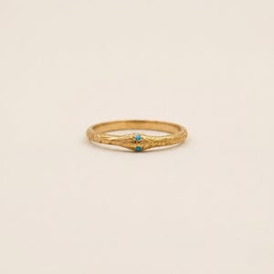 May include: A gold ring featuring an engraved snake-like pattern along the band. The head of the snake is detailed with two small, round, bright turquoise stones set as eyes, creating a distinct focal point on the textured metallic surface.