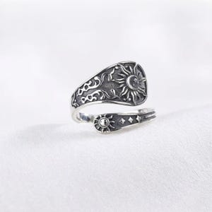 Puede incluir: Un anillo de plata oxidada con diseño abierto. El extremo más ancho muestra un motivo solar detallado con patrones de filigrana, mientras que el extremo más estrecho presenta un sol pequeño y símbolos de estrellas sobre un fondo oscuro y texturizado.
