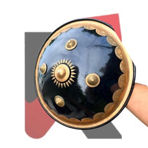 Pode incluir: Um escudo circular preto com uma borda dourada polida de acabamento festonado. O centro é adornado com um proeminente emblema solar dourado rodeado por quatro pequenos pinos cónicos dourados em relevo dispostos simetricamente.