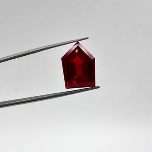 May include: A deep red, pentagon-shaped gemstone with a faceted surface held by a pair of thin, dark metal tweezers. The stone features a pointed top and a flat base, displaying a translucent, vibrant crimson color with sharp, geometric edges.