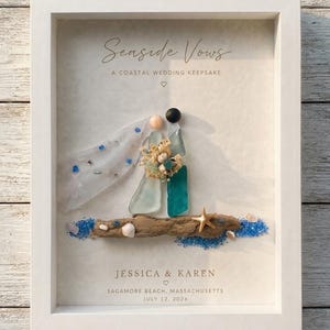 Może przedstawiać: Oprawiona dekoracja ścienna zatytułowana Seaside Vows, A Coastal Wedding Keepsake, przedstawiająca dwie postacie wykonane ze szkła morskiego stojące na kawałku drewna wyrzuconego przez morze. Postacie trzymają bukiet suszonych kwiatów. Tekst zawiera JESSICA & KAREN, SAGAMORE BEACH, MASSACHUSETTS, JULY 12, 2026, z niebieskim piaskiem i rozgwiazdą.