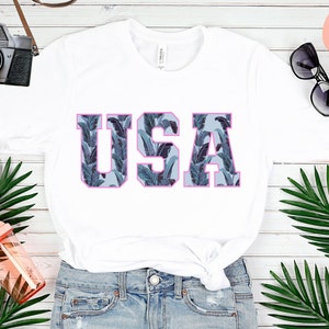 May include: White t-shirt with a graphic of the word "USA" in blue and white with a pink outline. The letters are filled with a tropical leaf pattern.