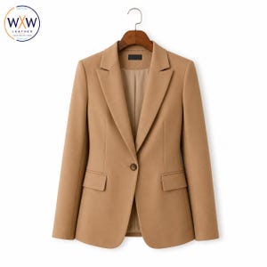 May include: A tan blazer featuring a single-button closure, notched lapels, and two front flap pockets. The garment is displayed on a wooden hanger and includes a small black interior label. The design is structured with long sleeves and a smooth finish.