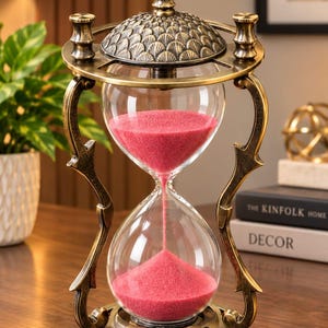 May include: An hourglass with a metallic bronze-toned frame featuring ornate, curved supports and a textured, scale-patterned top. The clear glass bulbs contain vibrant pink sand, with a thin stream currently flowing from the top to the bottom bulb.