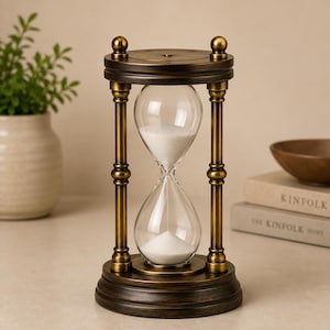 May include: An hourglass featuring a dark bronze-toned metal frame with a circular base and top, connected by four vertical pillars. The clear glass bulb contains white sand, with a portion of the sand currently resting in the bottom chamber.